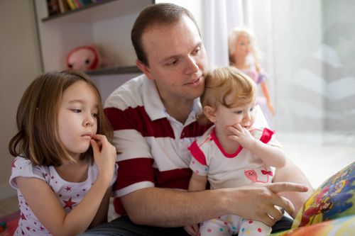 A man in a red and white striped shirt sits with his young daughters in their room and reads to them from a storybook.