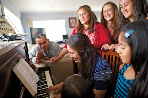 A young woman sits on a wooden chair, playing the piano as her sisters and father gather around singing.