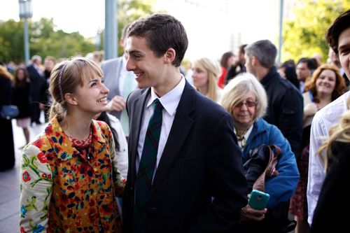 A smiling young man in a suit standing by a young woman in an orange jacket outside the Conference Center.
