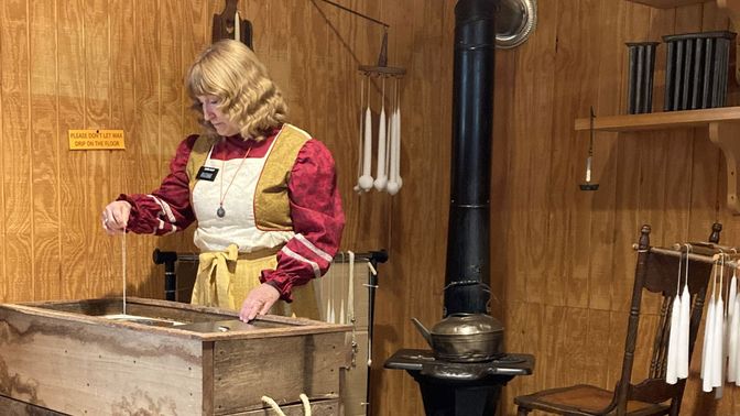 Interior showing missionary holding a string and looking into a tub. Iron stove and candles on display in the background.