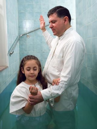 A girl with long brown hair, wearing a white jumpsuit, being baptized in a baptismal font by a man in a white shirt and tie.