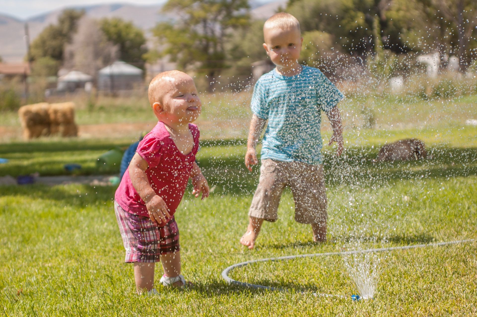 A baby and a toddler play in a sprinkler together outside.