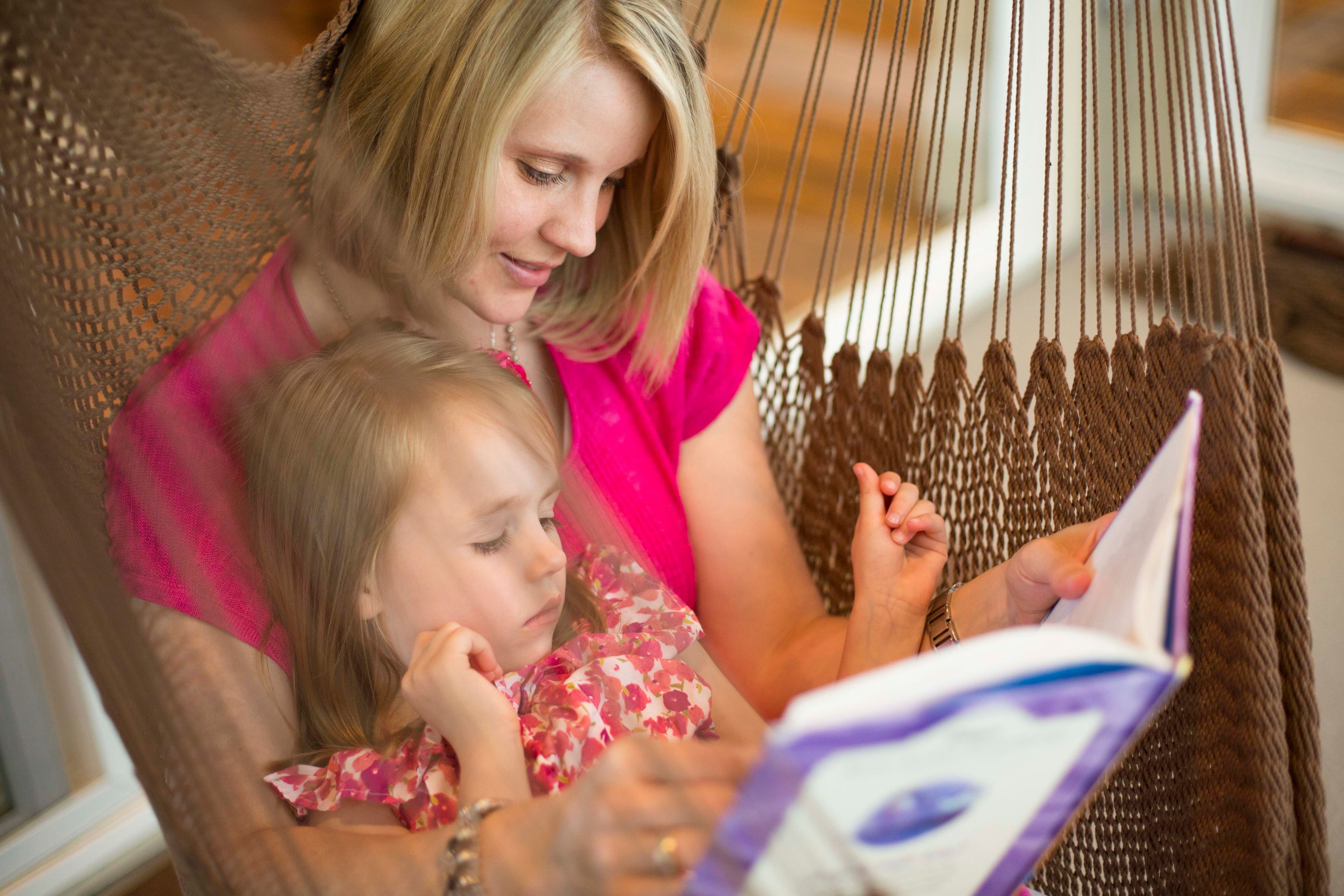 A mother and her daughter sit in a hammock together and read.