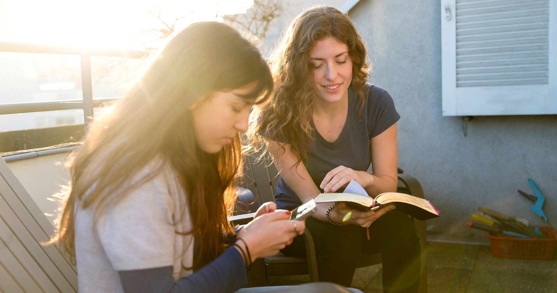 Two sister sit together on their balcony and read the scriptures together. One is reading from her smartphone and the other is reading from the paper version.