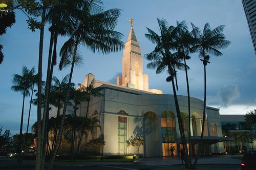 The entire Recife Brazil Temple lit up in the evening, with a view of the trees on the grounds of the temple and a partial view of city buildings nearby.