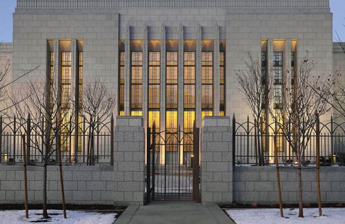 A gated fence on the grounds of the Draper Utah Temple in the evening, with the lighted windows of the temple seen beyond the fence.