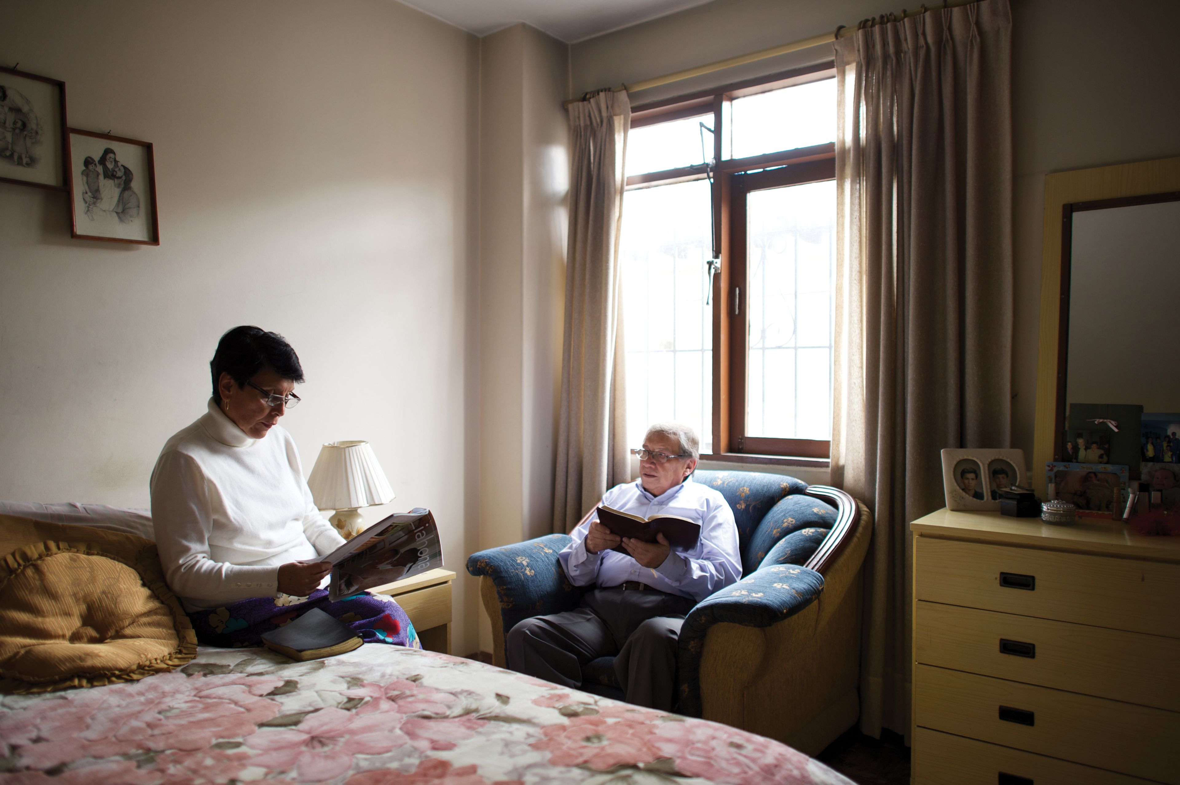 An elderly man reads the scriptures while his wife reads from a Liahona magazine in their bedroom.