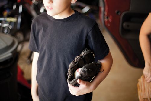 A boy in a black shirt holding a baseball and dark brown mitt under his arm.