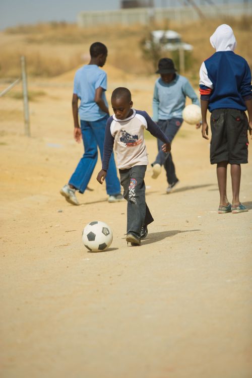 A little boy kicks a soccer ball on a dirt road with a group of other kids.