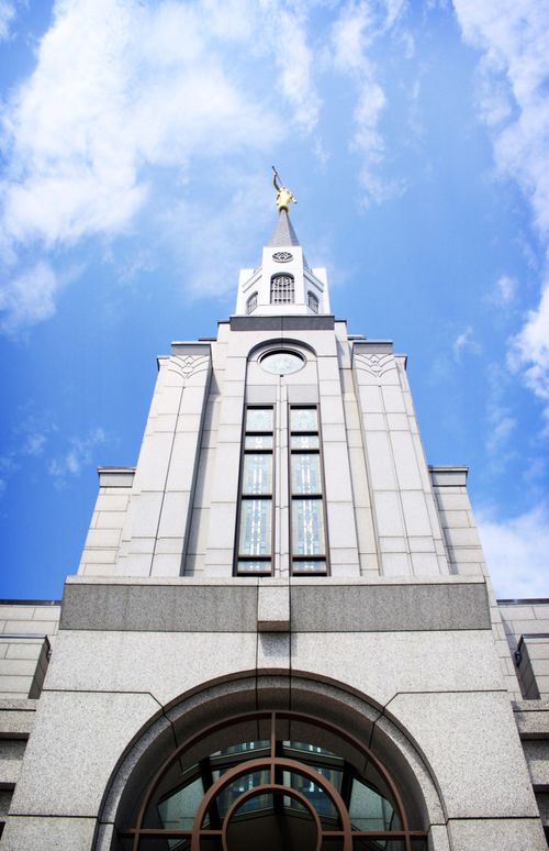 A view of the Boston Massachusetts Temple spire from below, with a blue sky and white clouds in the background.