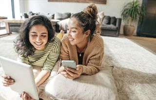 Two friends hanging out and using their digital devices. They are on their living room floor.