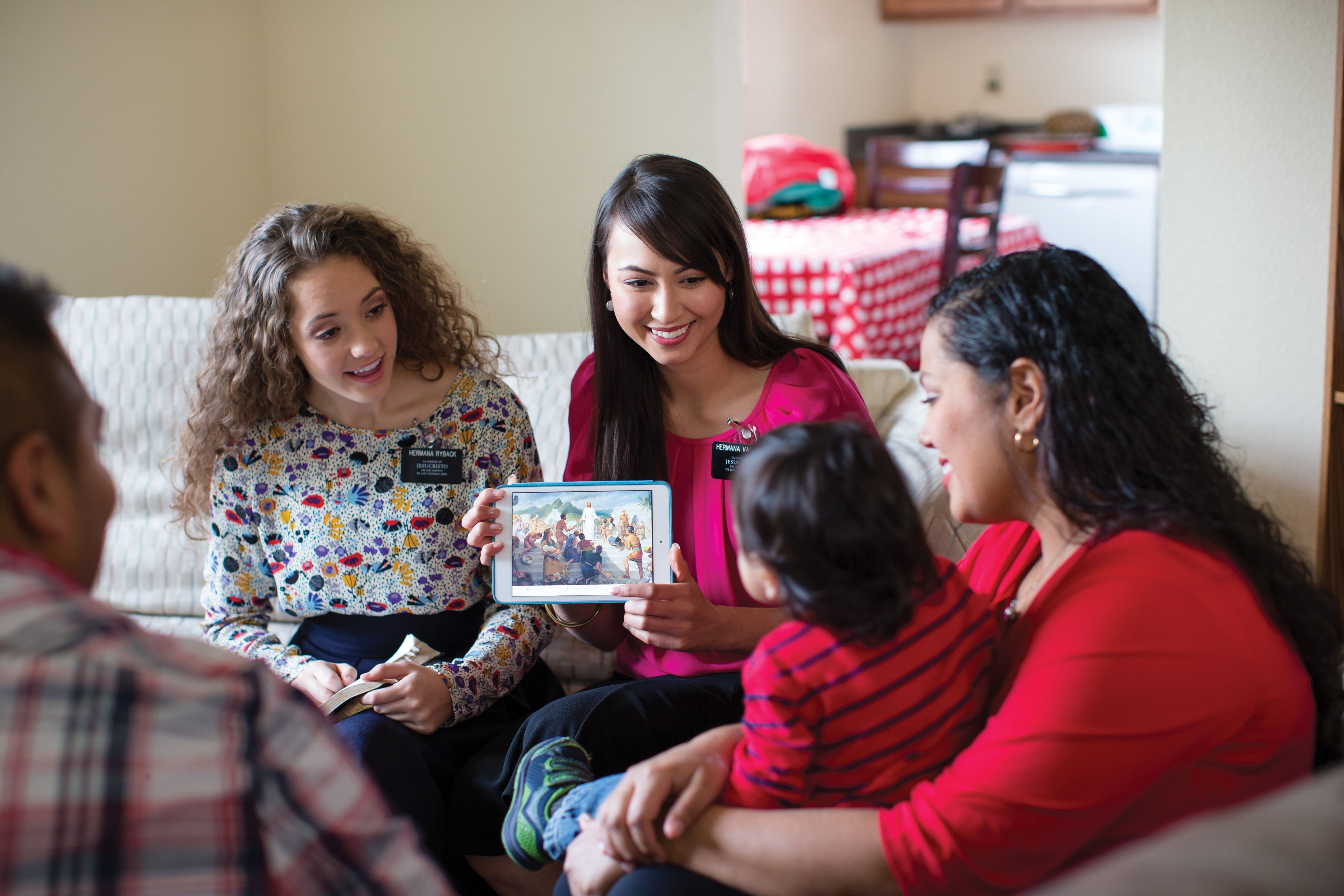 Two sister missionaries teach a family and show them gospel art on a tablet.