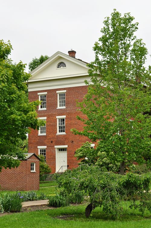 A red-brick building four stories tall with white doors and windows, surrounded by green grass, tall green trees, and shrubbery.