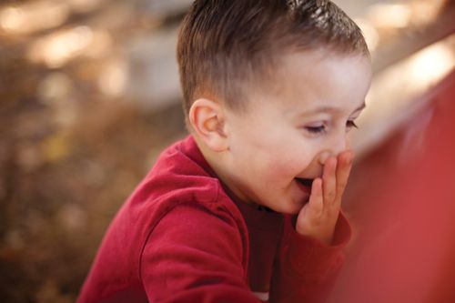 A little boy with short brown hair and a red shirt covers his mouth while laughing.