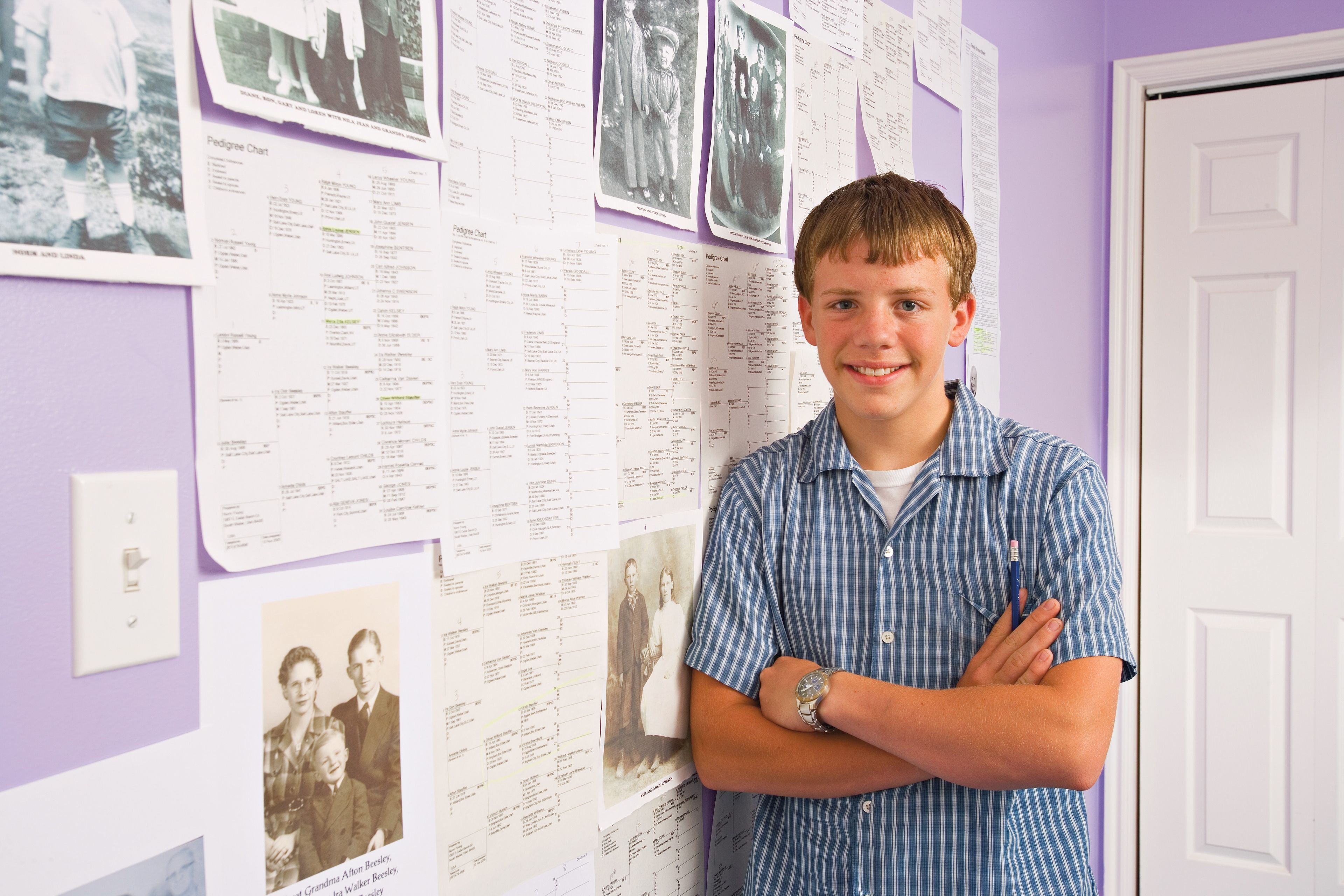 A young man stands against a wall covered in pedigree charts and photographs of his ancestors.