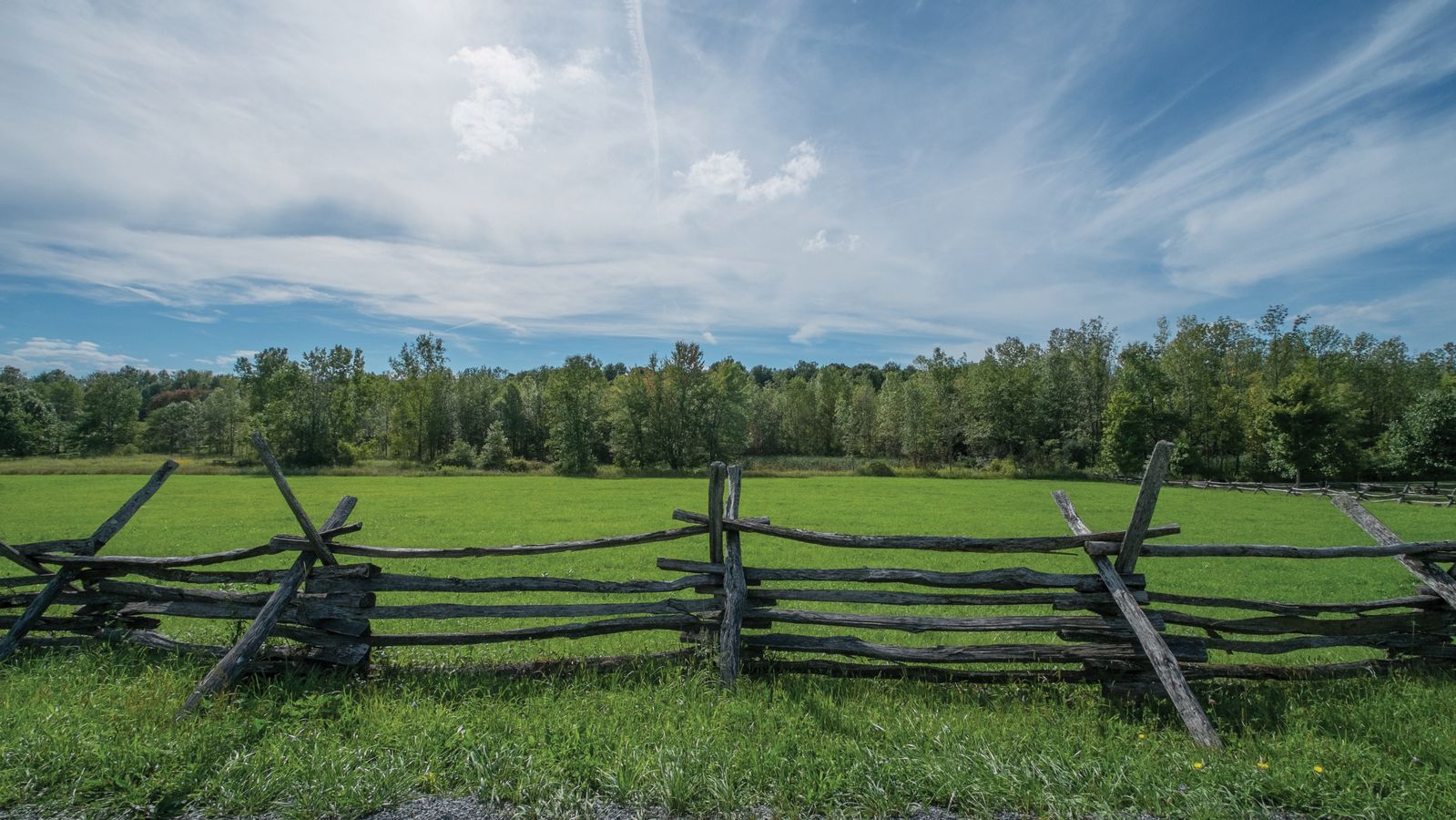 A wooden fence on the Smith family farm, running along the edge of a gravel road, with a green field and trees in the background.