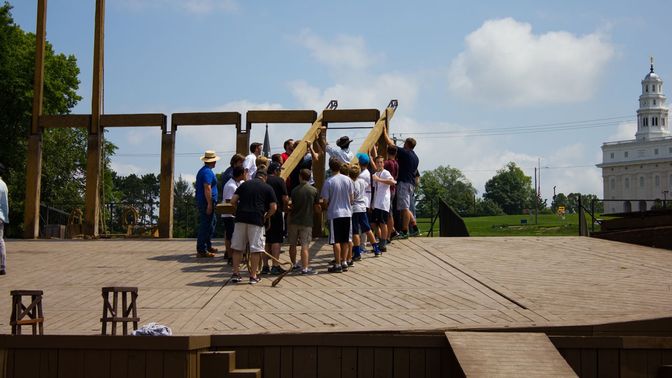 A group of men and young boys lifting large wooden beams on a stage to fit into a set piece at the back of the stage.