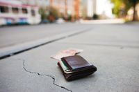 A man’s leather wallet lying on a cracked sidewalk, with a bill of money in front of it on the concrete and a city train blurred in the background.