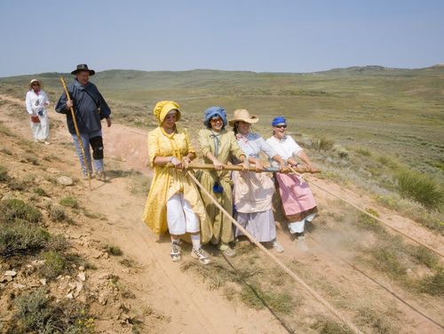 Four young women all dressed in hats, bonnets, and dresses pull back on a beam attached by ropes to a handcart as it goes downhill.
