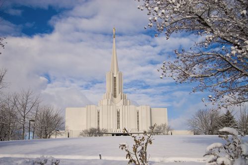 The Jordan River Utah Temple on a winter day, with a bare tree to the right side and a blue sky full of white clouds.