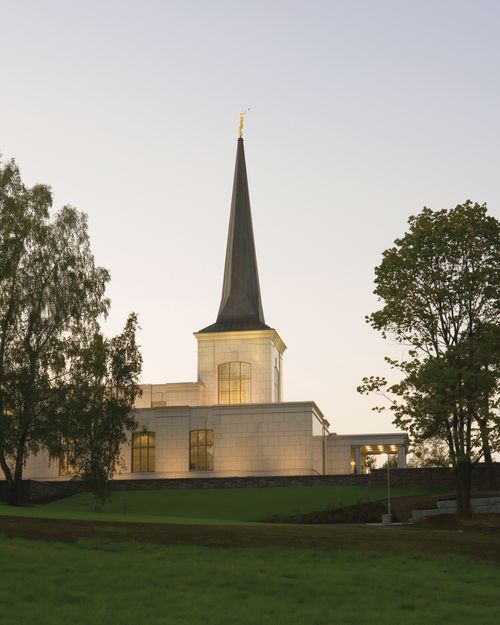 A side view of the spire on the Helsinki Finland Temple, with the lights on in the early evening and two large trees on either side.