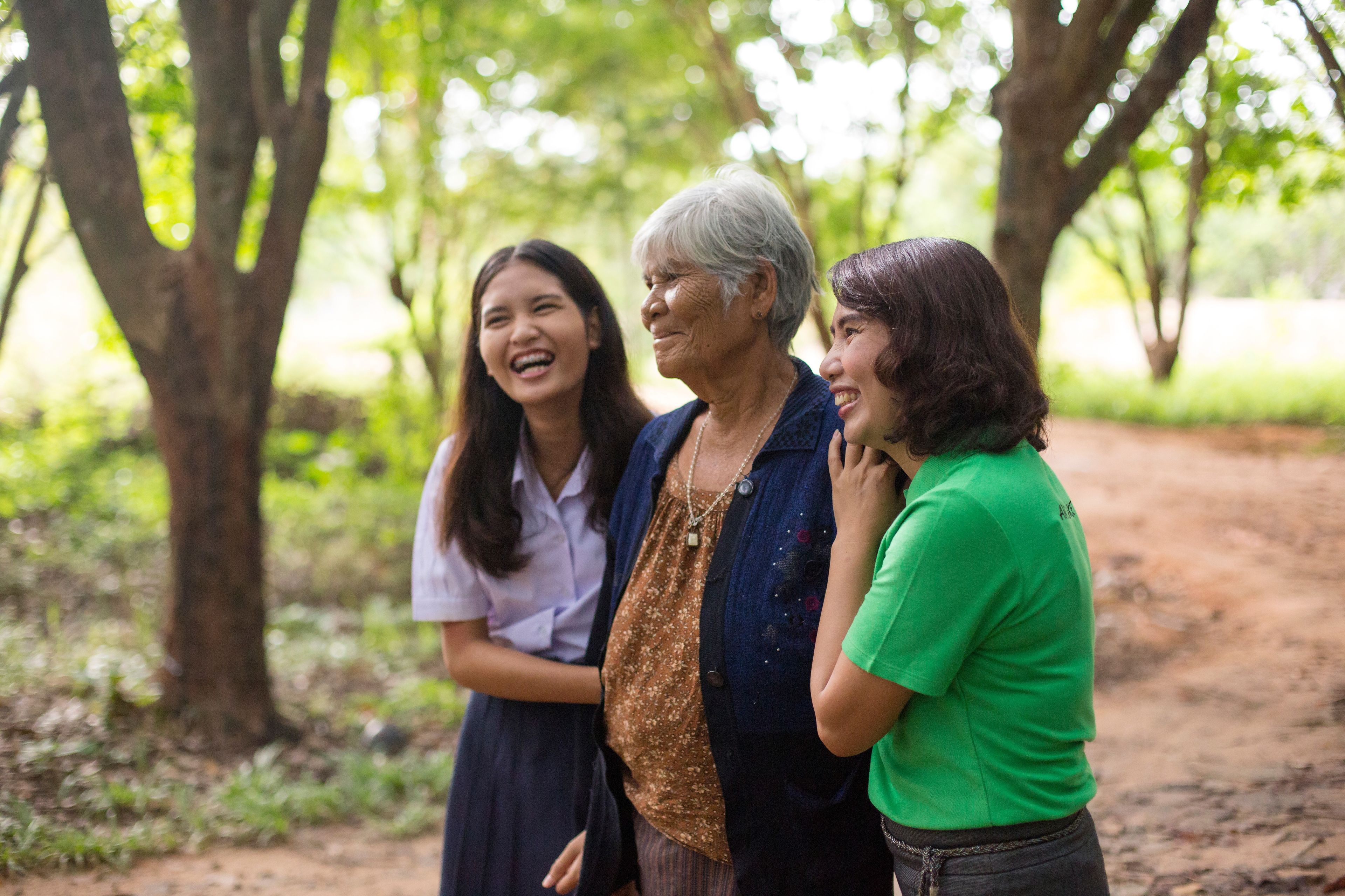 Two young women walk on a path through trees with an elderly woman.