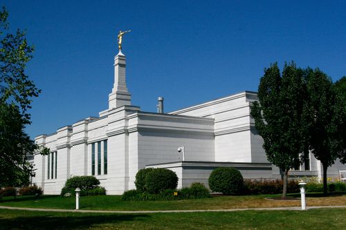 A view of the Palmyra New York Temple from one of the corners of the temple, with dark green trees in the foreground and a deep blue sky overhead.