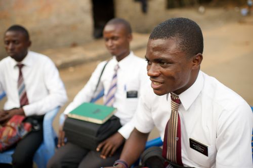 An elder missionary in Africa smiles while sitting outside at zone conference, with two other elder missionaries sitting behind him.