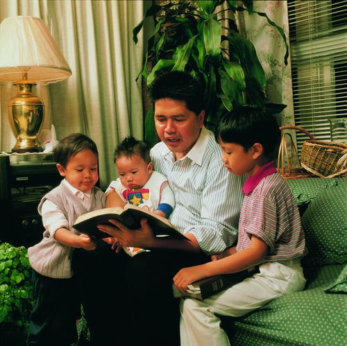 A father sitting on a sofa as he reads from the scriptures to two young boys and an infant held on his lap.