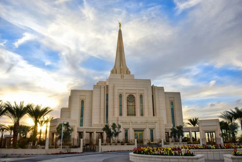 A view of the front entrance to the Gilbert Arizona Temple, with large white clouds overhead and the sun just beginning to set in the background.