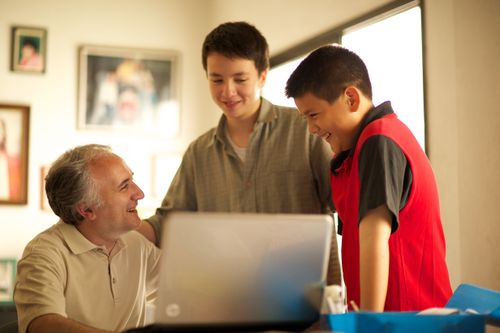 A father sits in front of his laptop at a table while his two sons stand next to him, one looking at the monitor and the other looking at the father.