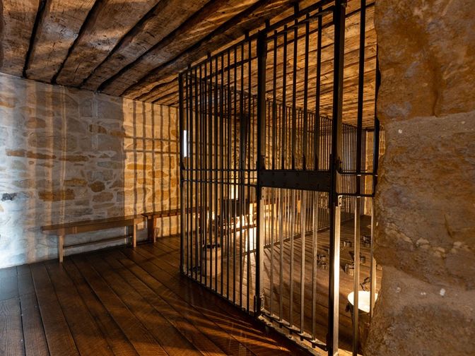 Small room with stone walls and a floor-to-ceiling metal cage. Light from a lantern illuminates the room. 