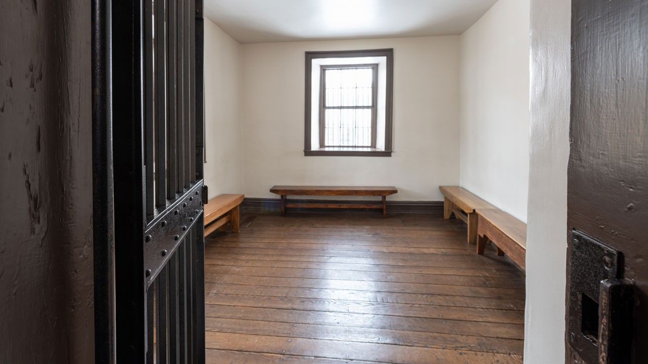 A metal bar door opens into a simple room with white walls, wood floors, a window, and benches.