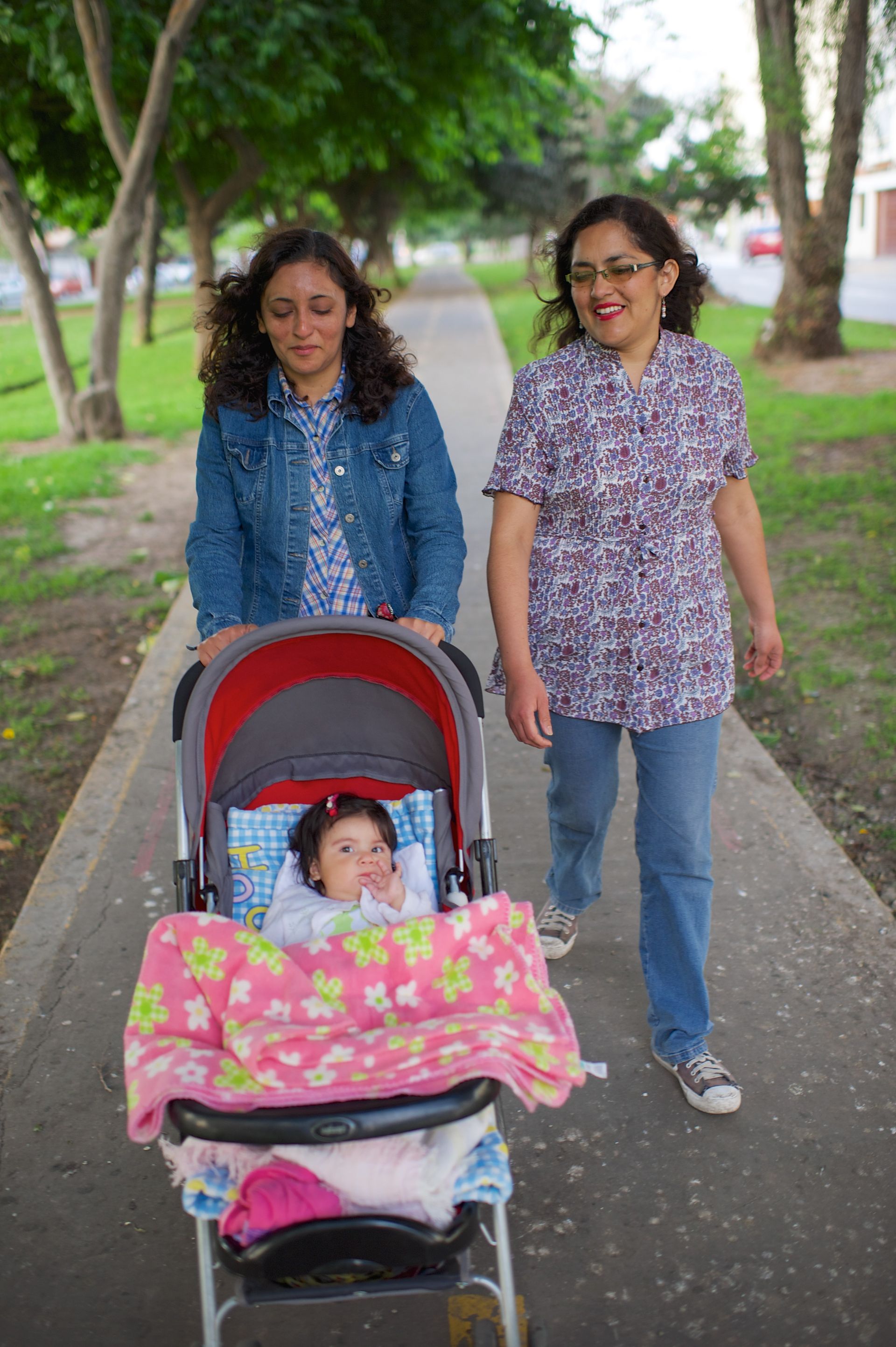 Two women push a baby in a stroller through a park.