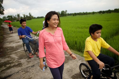 A woman in a pink shirt walks near her two sons, who are riding bicycles on a path surrounded by green fields.