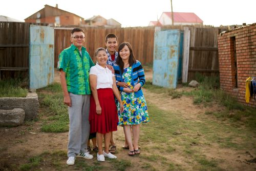 A father in Mongolia and his three adult children posing together outside, with a wooden fence and houses in the background.