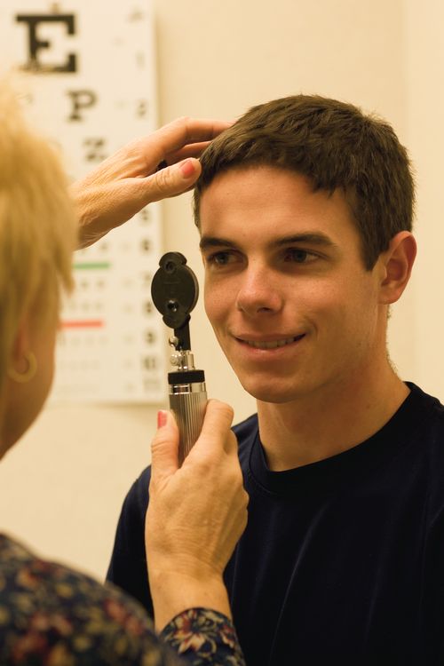 A dark-haired young man in a black shirt having his eyes checked by a doctor with a long metal device.