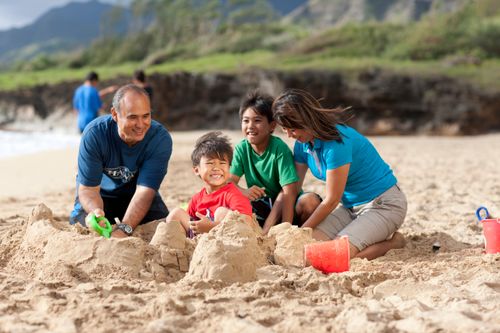 A mother and father kneel down in the sand and help their two young sons build a sand castle.