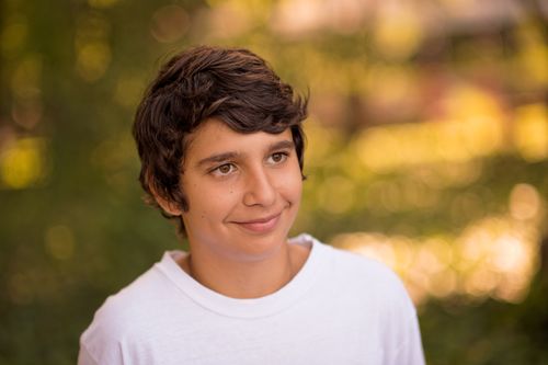 A portrait of a young man with brown wavy hair and a white T-shirt standing outside, looking to the side and smiling.