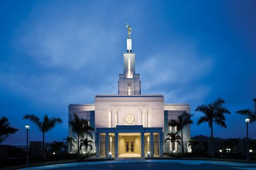 The front of the Panama City Panama Temple, with the lights on in the evening and palm trees growing on the path leading to the temple.