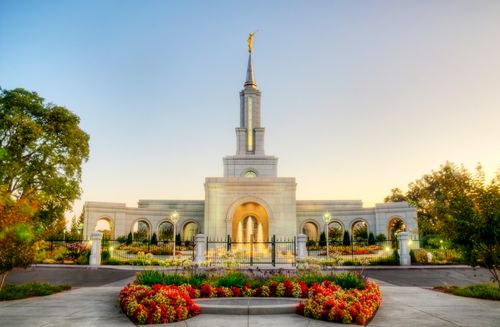 The Sacramento California Temple lit up in the early evening, with its fence, fountains, flowers, and trees.
