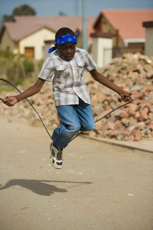 A boy in a plaid shirt and a blue bandanna plays jump rope down a street.