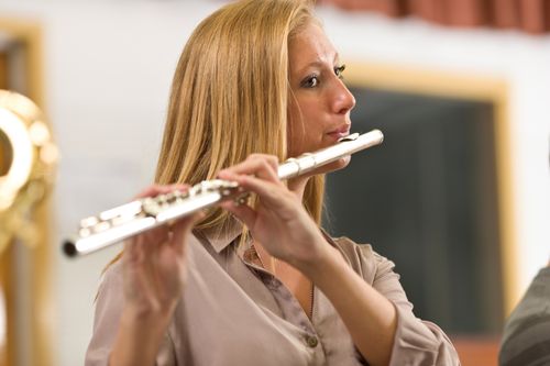 A young woman with long blonde hair stands and plays the flute with people on either side of her playing as well.