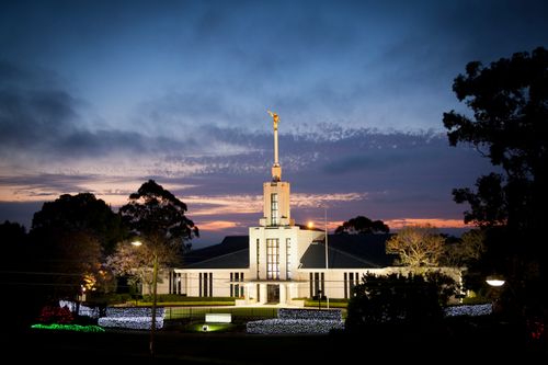 The entire Sydney Australia Temple, including the entrance, the spire, and the angel Moroni on top, with a fence surrounding the grounds, all lit up at night.