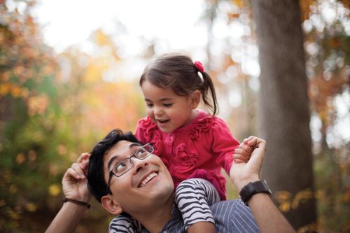 A father with glasses holds his daughter’s hands and smiles up at her as she sits on his shoulders.