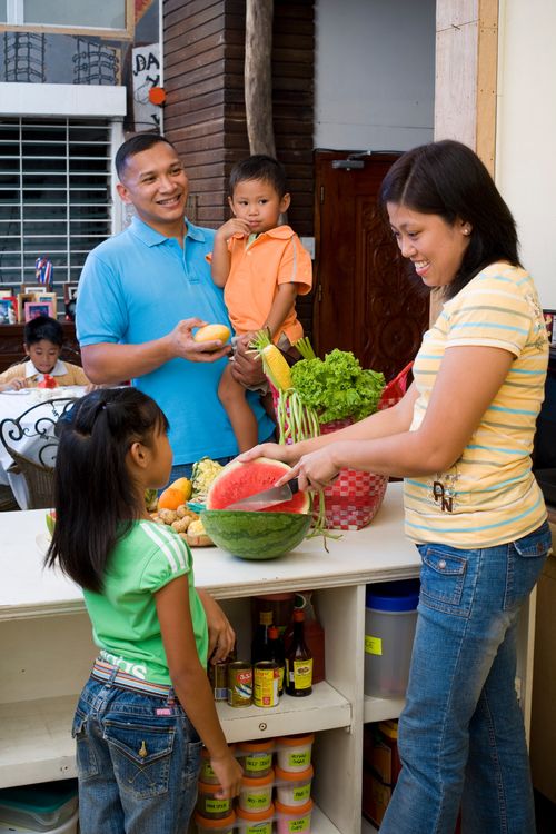 A mother and father cut up fruit in the kitchen while talking with their young children.