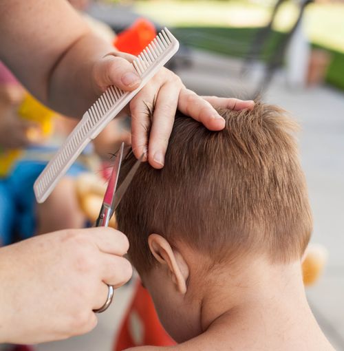 A woman’s hands are seen cutting a young boy’s hair with a comb and scissors.