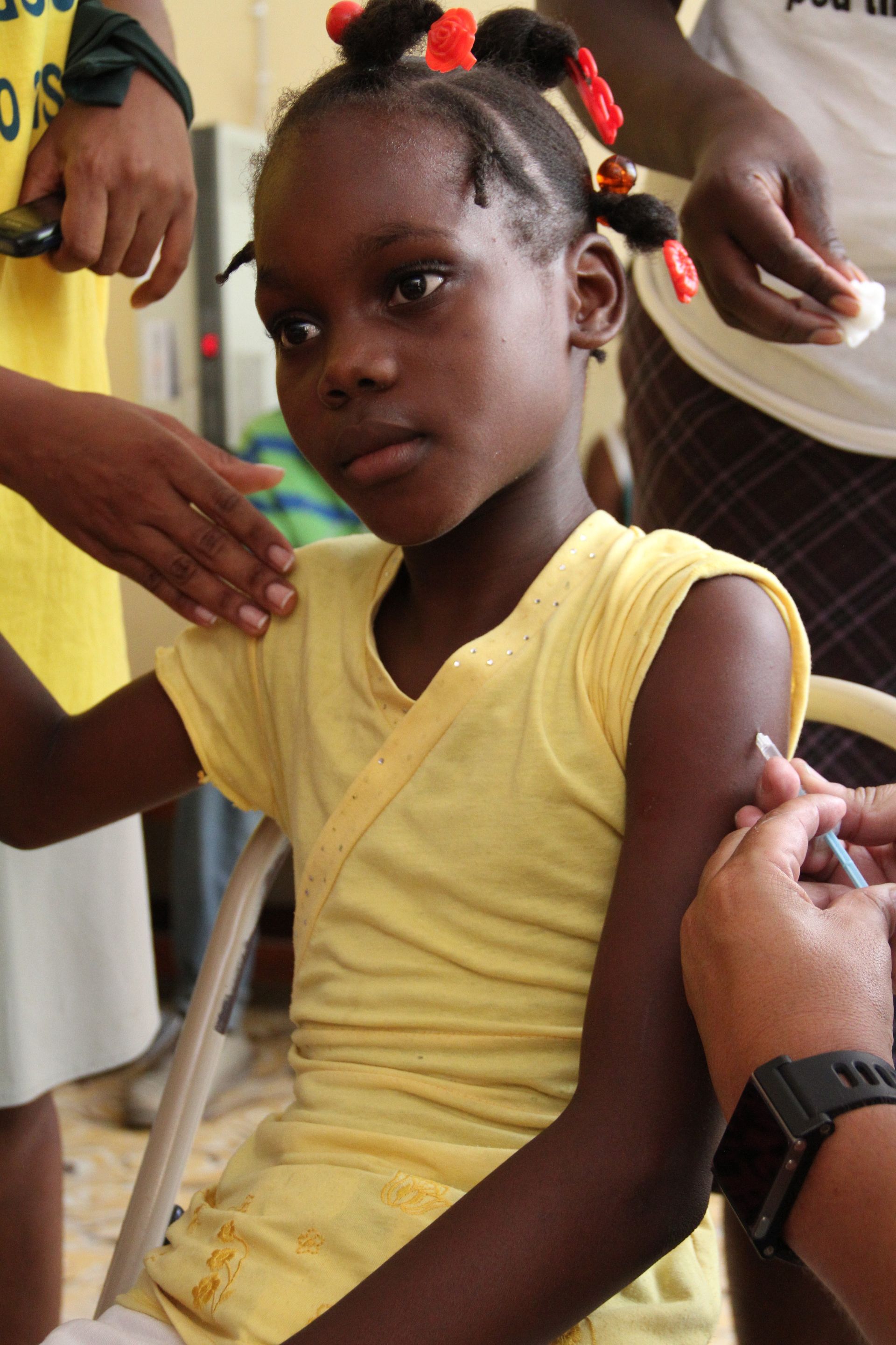 A young Haitian girl sitting down and receiving an immunization in her arm.