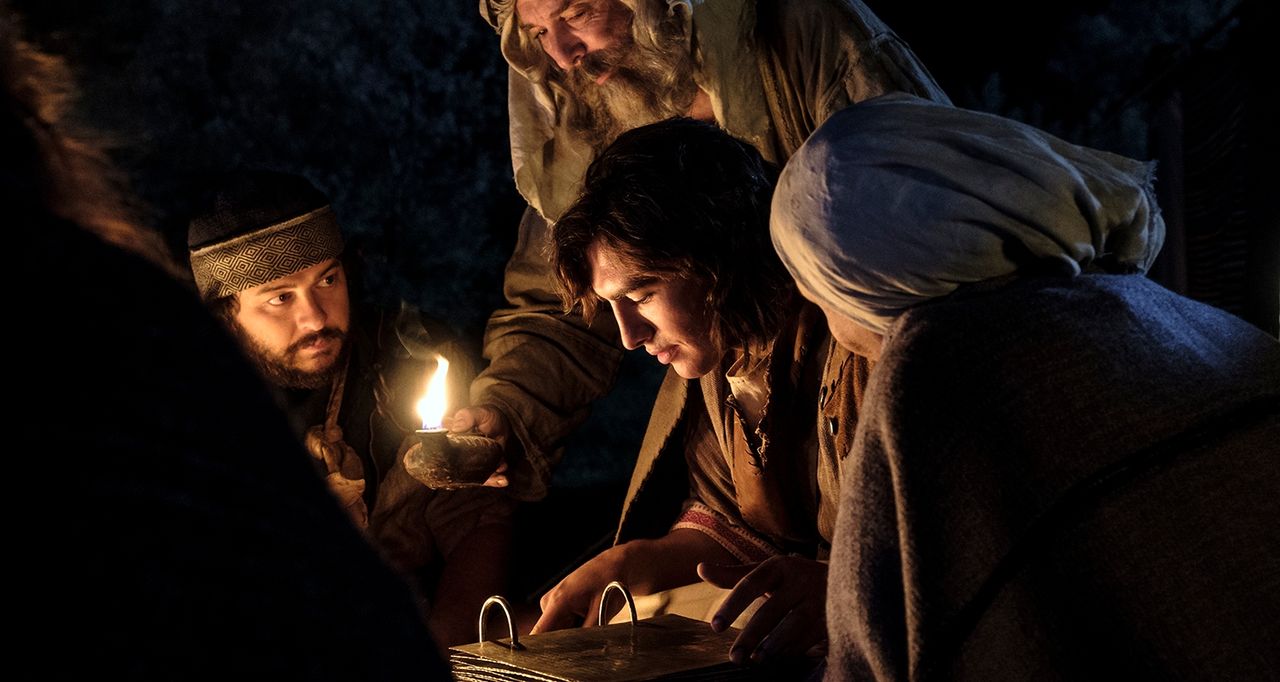 The Brass Plates are being held by Nephi. They are being viewed through an oil lamp. Lehi, Zoram, and Sariah all stand near Nephi looking at the plates. This is part of the Book of Mormon Visual Library. 

This is from Scene 232: Lehi studies the brass plates