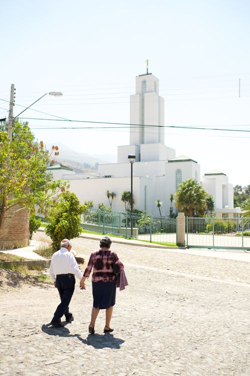 An elderly couple holding hands and walking across a street in front of the Cochabamba Bolivia Temple.